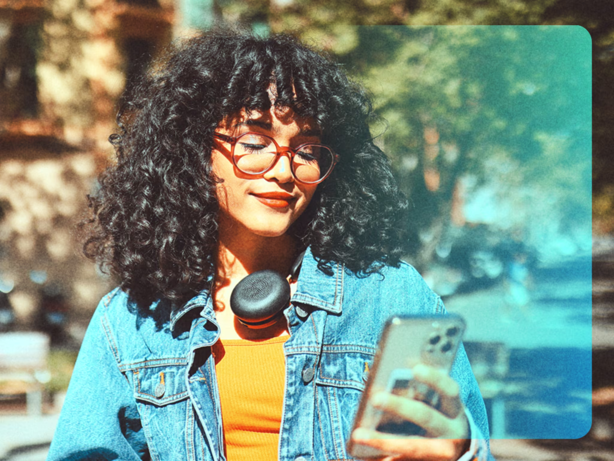 Junge Frau mit Kopfhörer um den Hals blickt auf ihr Smartphone, im Hintergrund eine sonnige Stadtstraße.