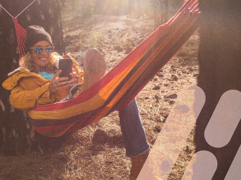 Frau liegt entspannt in einer Hängematte und schaut lächelnd auf ihr Smartphone.