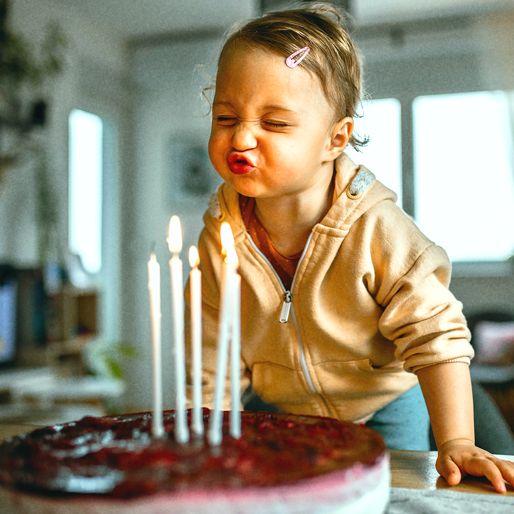 Ein kleines Kind mit blondem Haar und Haarspange pustet mit geschlossenen Augen und spitzem Mund konzentriert die Kerzen auf einem Geburtstagskuchen aus. Fünf brennende Kerzen stehen auf einem dunklen, roten Kuchen. Die Szene wirkt lebendig und liebevoll.