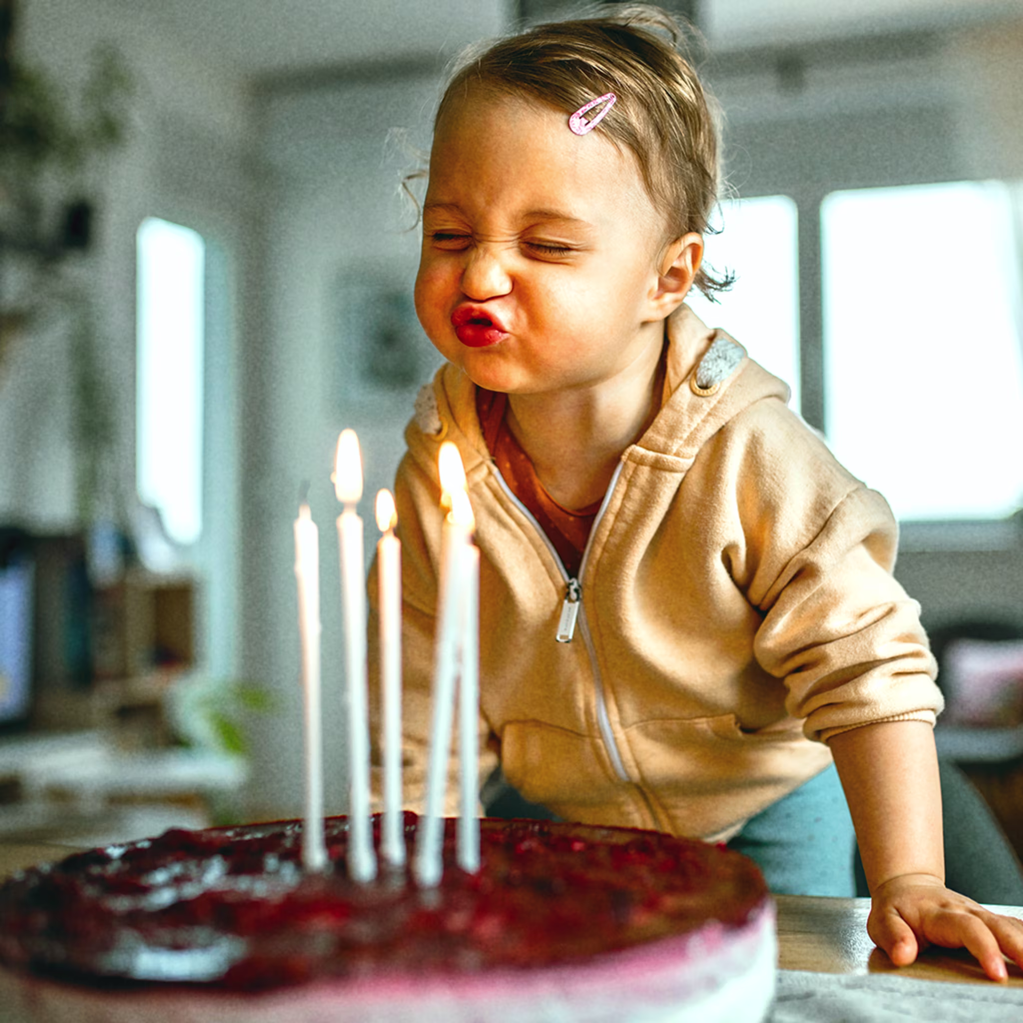 Ein kleines Kind mit blondem Haar und Haarspange pustet mit geschlossenen Augen und spitzem Mund konzentriert die Kerzen auf einem Geburtstagskuchen aus. Fünf brennende Kerzen stehen auf einem dunklen, roten Kuchen. Die Szene wirkt lebendig und liebevoll.