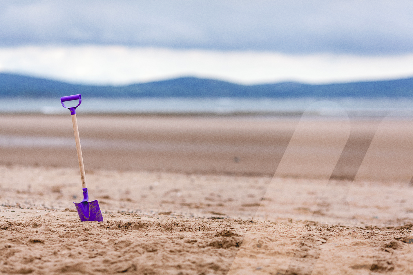 Lila Kinderschaufel mit Holzstiel steckt im Sand eines menschenleeren Strandes. Im Hintergrund sind das Meer und eine verschwommene Berglandschaft unter bewölktem Himmel zu sehen.