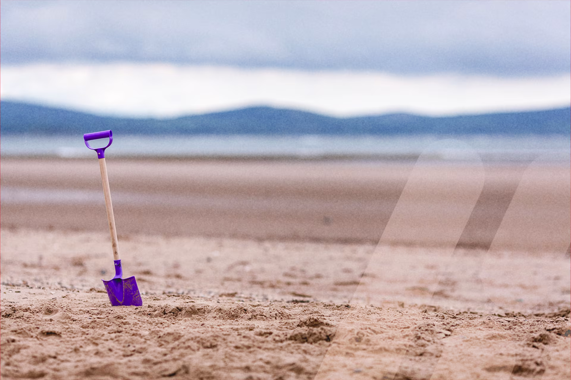 Lila Kinderschaufel mit Holzstiel steckt im Sand eines menschenleeren Strandes. Im Hintergrund sind das Meer und eine verschwommene Berglandschaft unter bewölktem Himmel zu sehen.