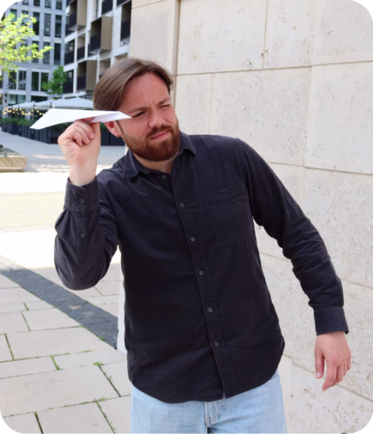 Ein Mann mit Bart und dunklem Hemd hält ein gefaltetes Papierflugzeug in Wurfposition und schaut konzentriert nach vorne. Er steht im Freien vor einem modernen Gebäude.
