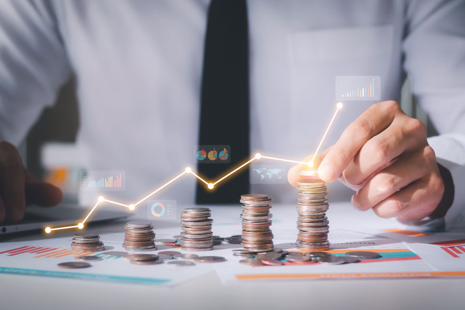 Geschäftsperson stapelt Münzen auf einem Tisch; die Stapel werden höher. Darüber verläuft eine steigende Linie mit Diagramm-Icons – Symbol für Wachstum, Vermögensaufbau und finanzielle Entwicklung.
