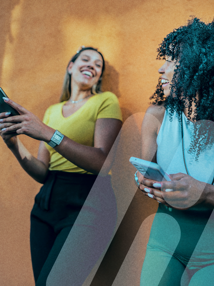 Zwei junge Frauen lachen miteinander und schauen sich an, während sie ihre Smartphones halten, vor einer sonnig-orangefarbenen Wand