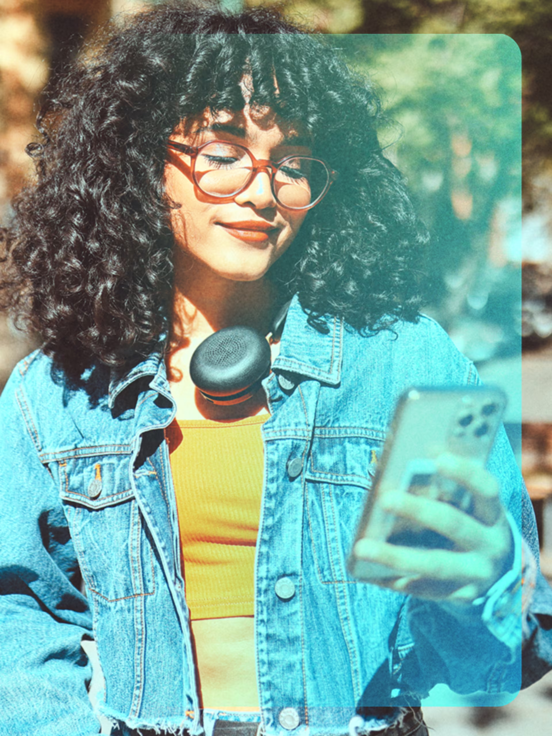 Junge Frau mit lockigen Haaren, Brille und Kopfhörern um den Hals steht draußen an einem sonnigen Tag und schaut lächelnd auf ihr Smartphone. Sie trägt eine Jeansjacke über einem orangefarbenen Oberteil.  Die Szene wirkt entspannt und urban