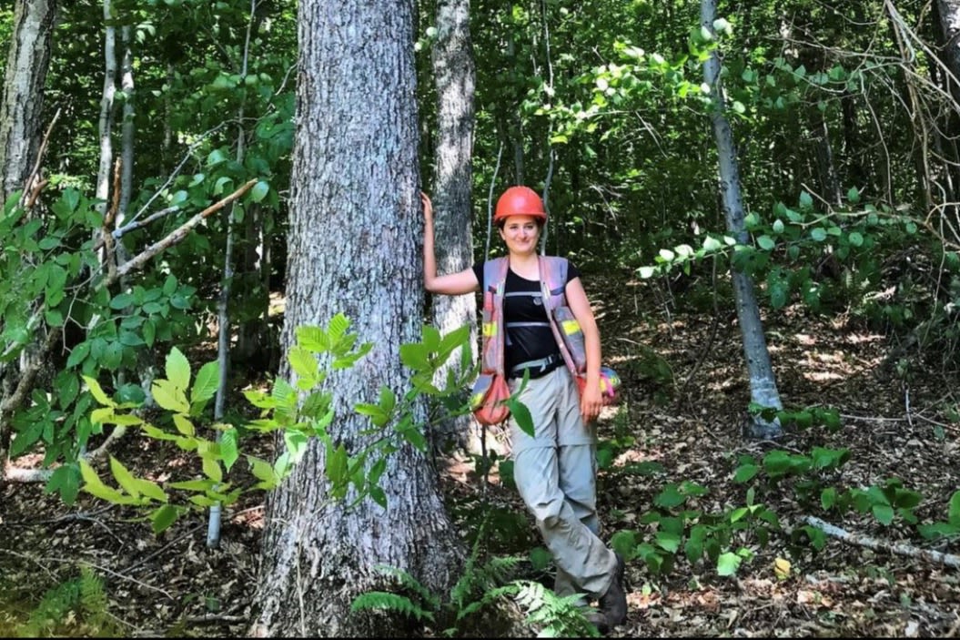 Sierra Giraud stands in a green forest, leaning on a tree in full forestry gear.