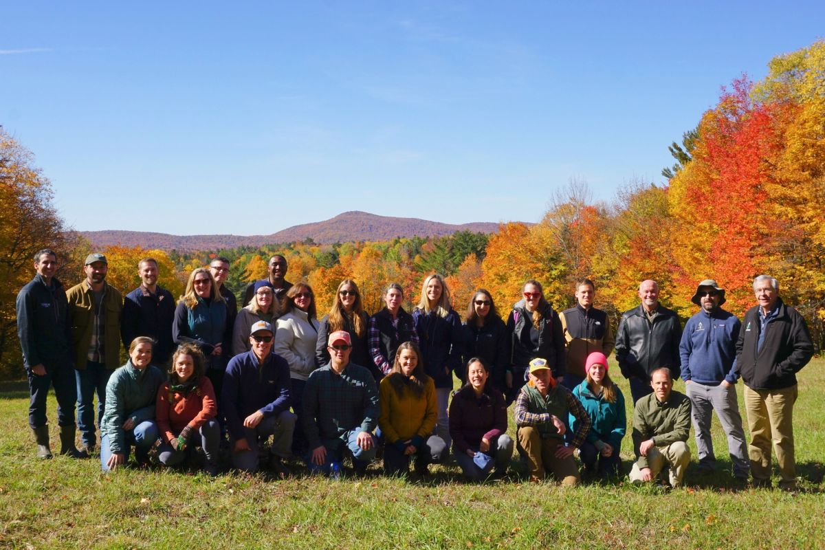 A large group of people pose outside for a photo with mountains and vibrant fall foliage behind them.