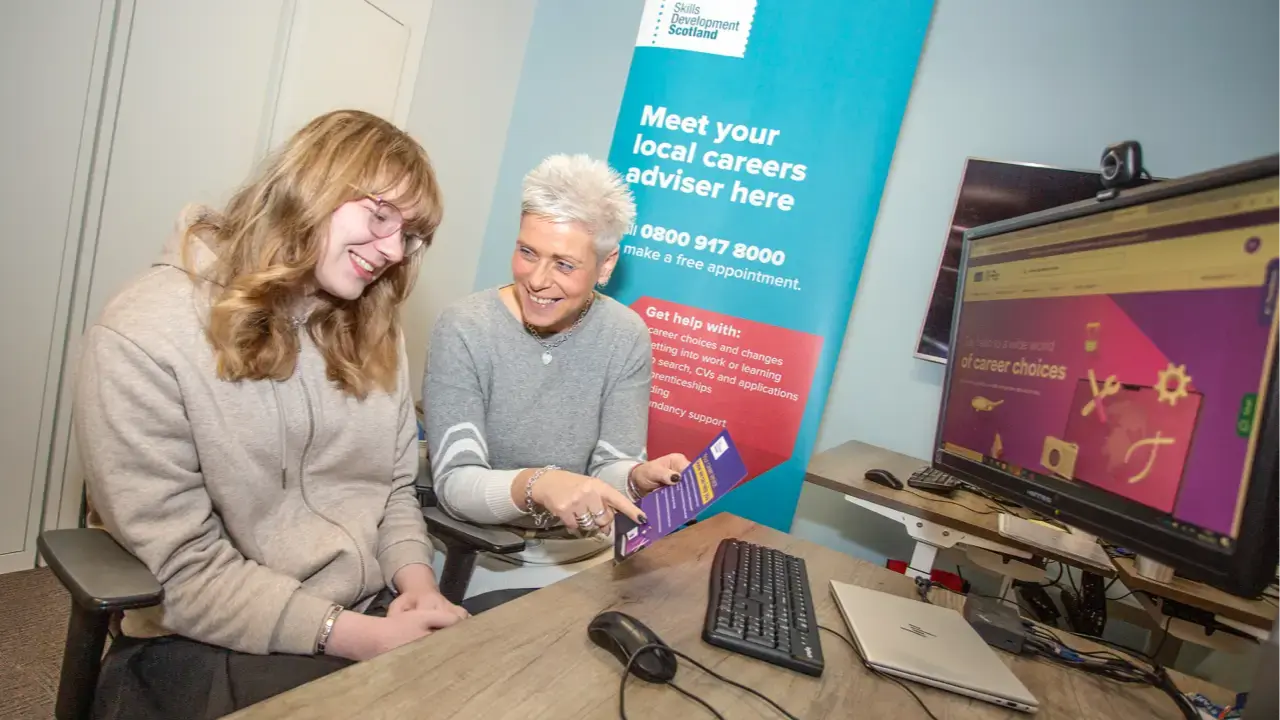 Cloe (on left) smiling at leaflet in front of computer sitting next to Emma (on right) who is holding the leaflet