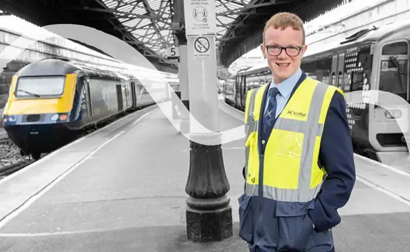 Smiling young man wearing ScotRail uniform, standing beside trains on platform