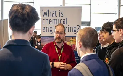 A man talks to some school pupils. A sign in the background says 'what is chemical engineering?'
