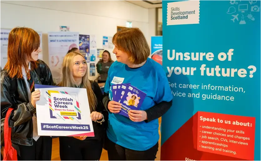 Two school girls talk to a Skills Development Scotland careers adviser. One of the girls holds a promotional Scottish Careers Week sign.
