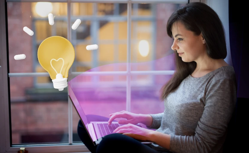 A person sitting by a window on their laptop, there's an illustration of a light bulb above the laptop.