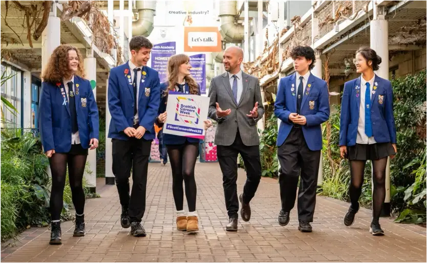 A group of four school pupils walk alongside a man and a woman. Each school pupil is wearing a blue blazer and the adult woman is holding a promotional Scottish Careers Week sign.