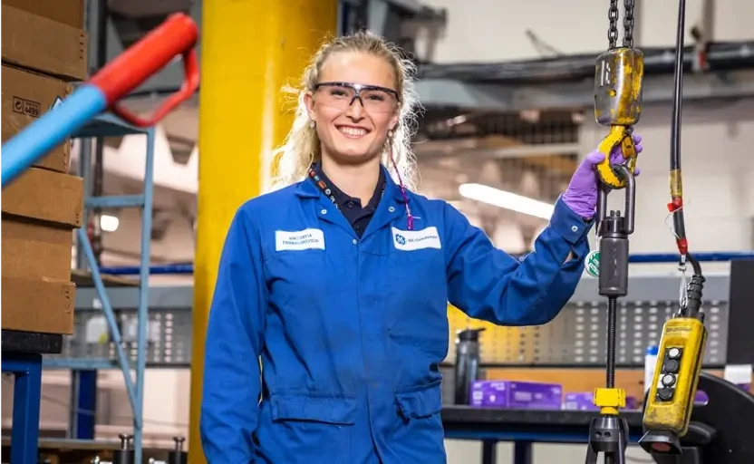 A young apprentice in a workshop smiling, they're wearing blue work overalls and safety glasses