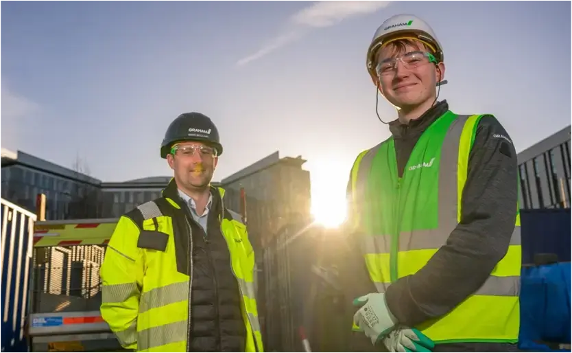 Two men wearing hi-vis jackets and hard hats stand on a building site.