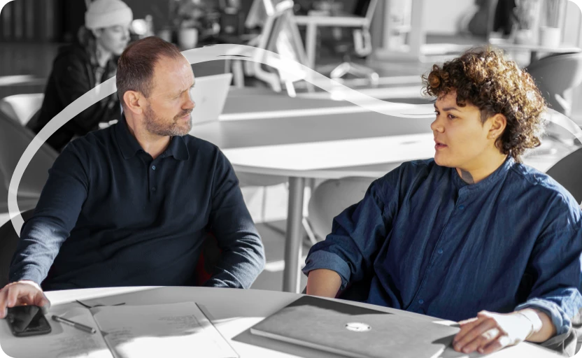 A young person sitting at a desk with a laptop, they're having a conversation with an older person next to them.