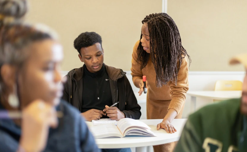 A young learner sitting at a desk over a textbook, getting instruction from a teacher.