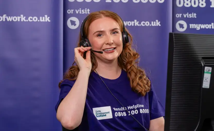 A woman, wearing a purple Results Helpline t-shirt, sits at a desk while holding her headset.