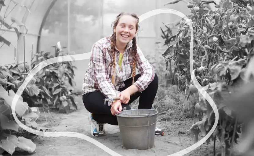 A young person working in a greenhouse. They're crouching among the plants, looking at the camera and smiling.