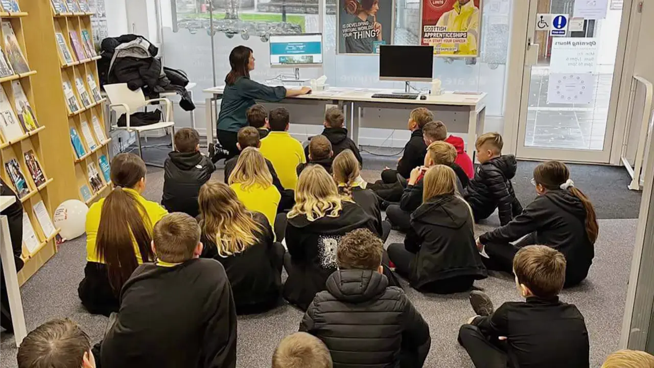 A teacher sitting down with a class of pupils, showing them something on a computer