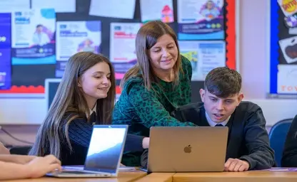 A woman shows stands behind two school pupils and shows them something on a laptop screen.