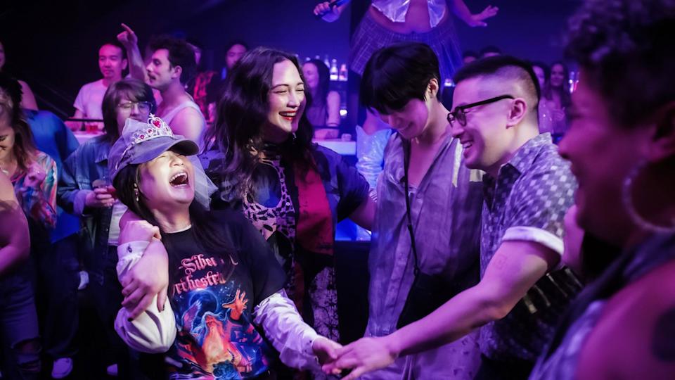 Diverse group dancing at nightclub with purple lighting; person wearing tiara and graphic t-shirt laughing joyfully.