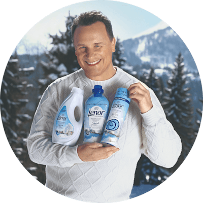 Person in einem weißen Pullover hält drei Lenor Winterzauber Produkte vor einer verschneiten Berglandschaft mit Tannenbäumen im Hintergrund.