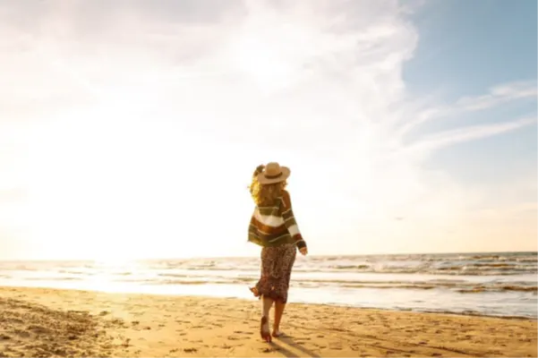 Frau läuft barfuß am Strand entlang und blickt auf das Meer bei warmem Sonnenuntergang, sanfte Wellen und weiter Himmel im Hintergrund