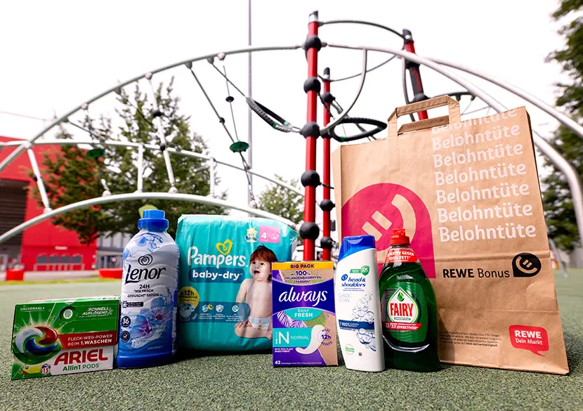 Verschiedene P&G Produkte und eine REWE-Papiertragetasche stehen nebeneinander auf dem Boden. Im Hintergrund ist ein inklusives Spiel- und Klettergerät auf einem Stück zum Glück Spielplatz zu sehen. 