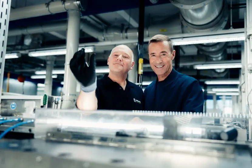 Foto von einem Gillette Mitarbeitenden und Kai Pflaume in einer Fabrikumgebung, wobei der Mann Kai eine Gillette Rasierklinge zeigt.