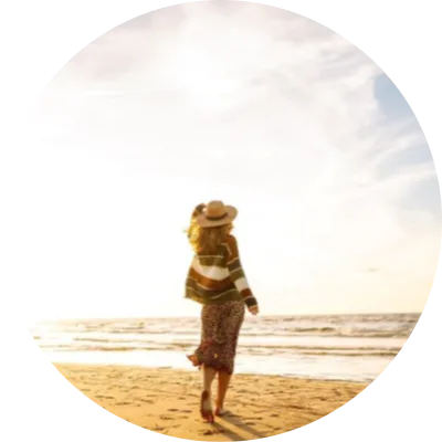 Frau läuft barfuß am Strand entlang und blickt auf das Meer bei warmem Sonnenuntergang, sanfte Wellen und weiter Himmel im Hintergrund