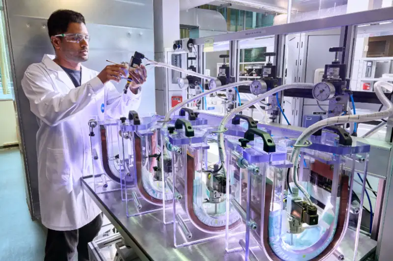 Foto eines Wissenschaftlers in einem Laborkittel und mit Schutzbrille, der an einem Metalltisch mit einer komplexen Versuchsanordnung aus durchsichtigen Rohren, Manometern und Ventilen arbeitet.