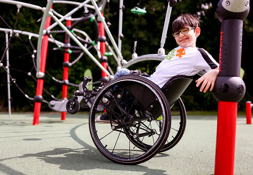 Ein Junge im Rollstuhl lehnt lächelnd und lässig nach hinten gelehnt an einem Spielgerät auf einem inklusiven Spielplatz