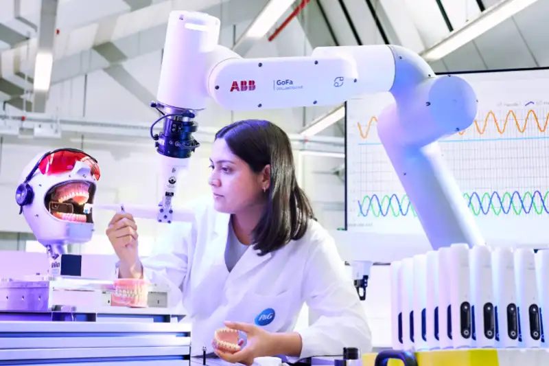 Foto einer Laborumgebung, in der eine Frau in einem weißen Laborkittel einen Roboterarm bedient, um mit einer zahnmedizinischen Übungspuppe zu interagieren.