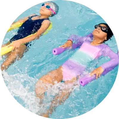 Zwei Kinder treiben beim Schwimmunterricht mit Schwimmnudeln und Schwimmbrillen entspannt auf dem Rücken im Schwimmbecken und üben das Rückenschwimmen.