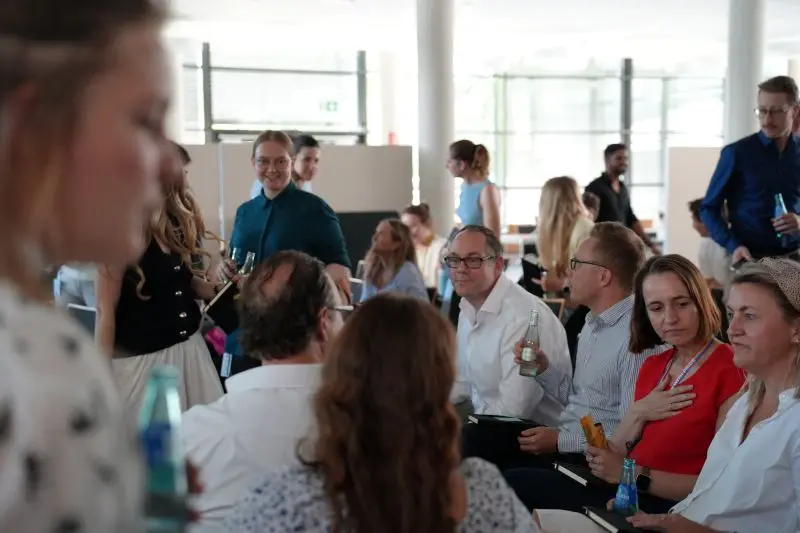 Mitarbeitende unterschiedlichen Alters in einem hellen Raum, die sich in kleinen Teams unterhalten und vernetzen