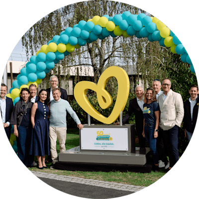 Gruppe von Personen um eine goldene Herzskulptur unter einem blau-gelben Ballonbogen zur Feier von 50 Jahren Pampers-Produktion in Euskirchen.