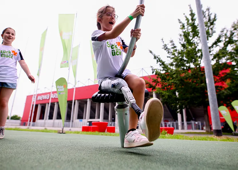 Ein junges Mädchen mit Beinprothese sitzt lachend und sich festhaltend auf einem Spielgerät auf einem inklusiven Spielplatz. Im Hintergrund sind ein weiteres Mädchen und grüne "Stück zum Glück" Fahnen zu sehen.  