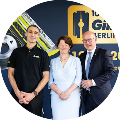 Drei Personen posieren vor einer Kulisse zum 100-jährigen Jubiläum von Gillette Berlin mit Gillette-Rasierprodukten und Markenlogo.