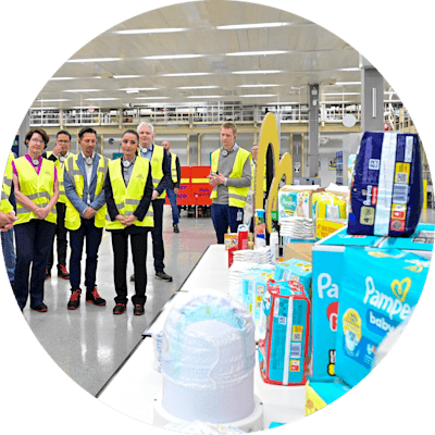 „Personengruppe mit Warnwesten bei einer Werksführung in einer Produktionshalle mit ausgestellten Konsumgütern wie Pampers‑Windeln.