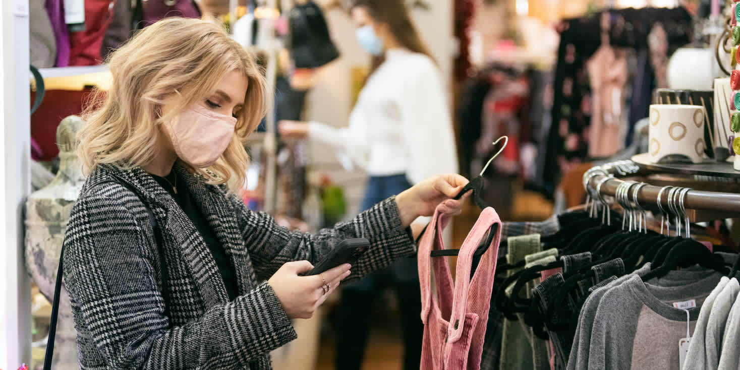 Woman shopper with mask looking at her phone while holding a pink garment in-store, making a decision.