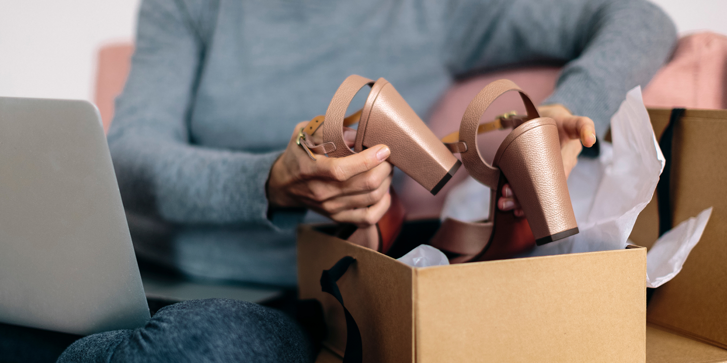 Woman opening box of high-heeled sandals she bought online, with open laptop on her lap.