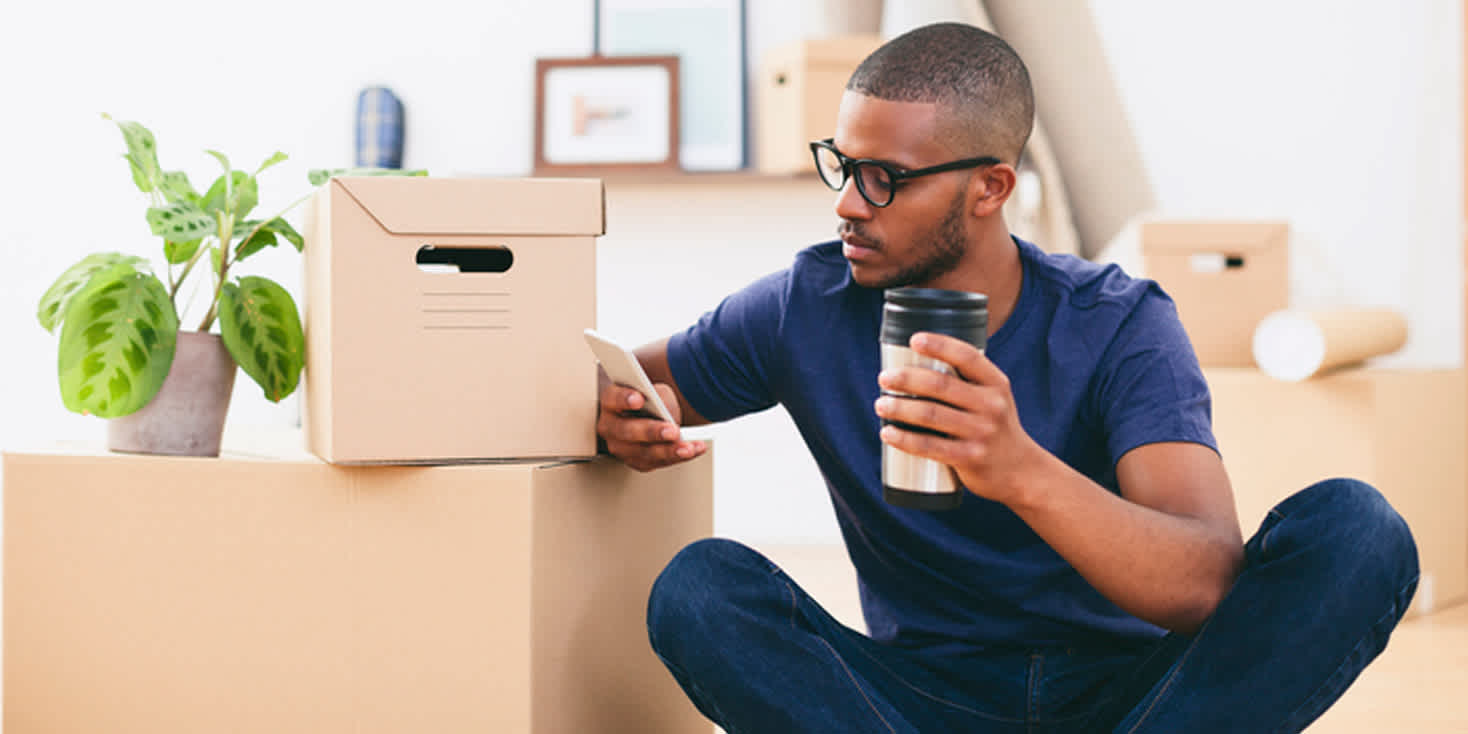 Young Black man sitting among moving boxes as he checks his phone and sips coffee.
