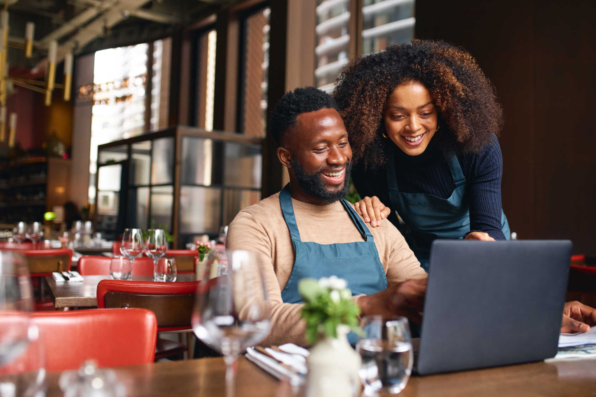 Male restaurant owner, smiling, reviewing figures on his laptop while female colleague looks on over his shoulder.