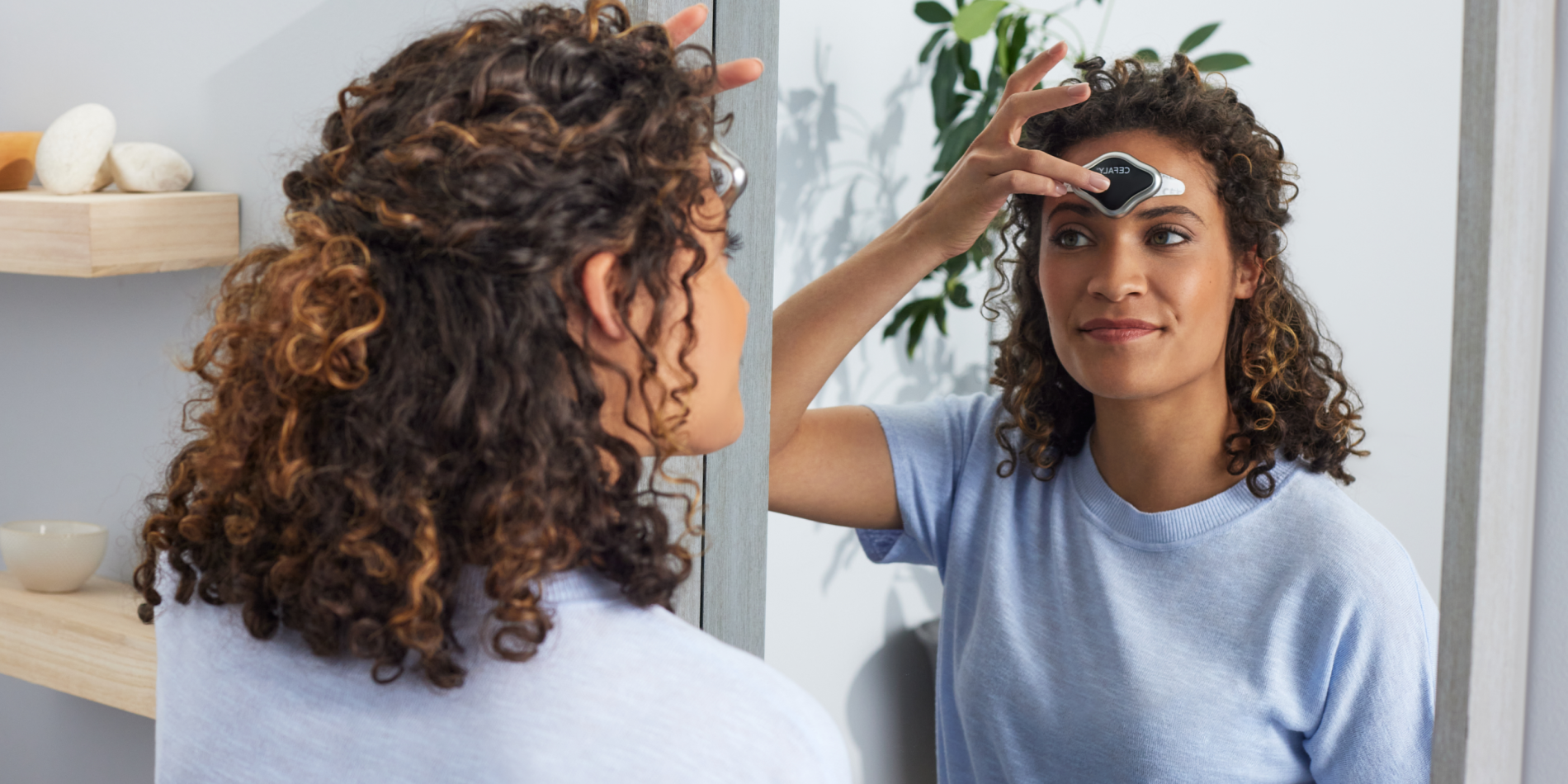 Young woman looking in mirror while holding a CEFALY device to her forehead