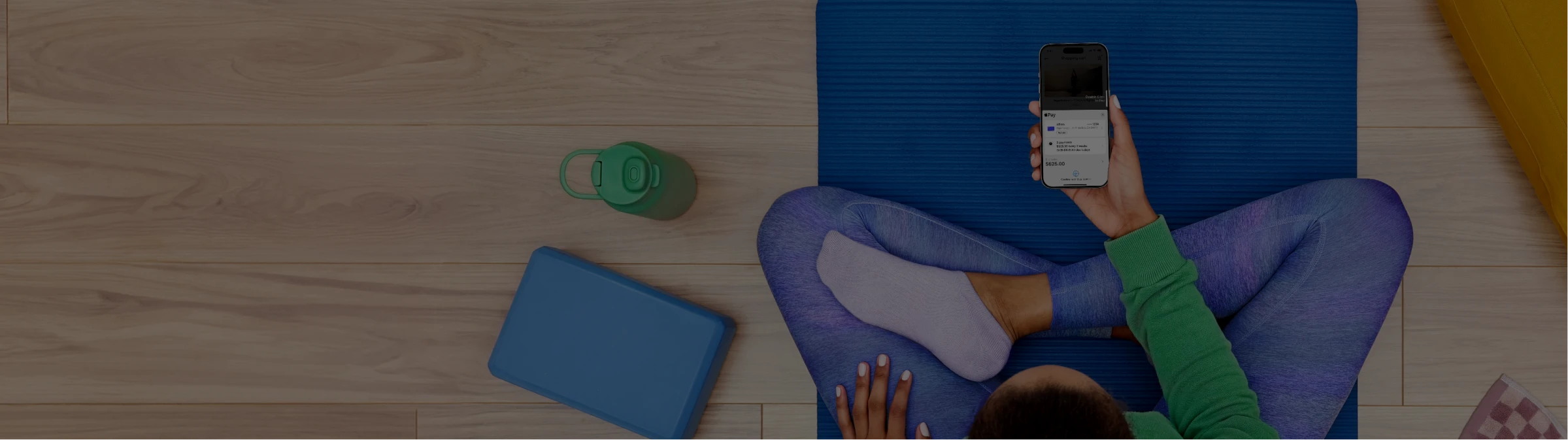 A person sitting cross-legged on a blue yoga mat holding a smartphone with a payment plan interface displayed, surrounded by a yoga bolster, water bottle, and other fitness accessories on a wooden floor.