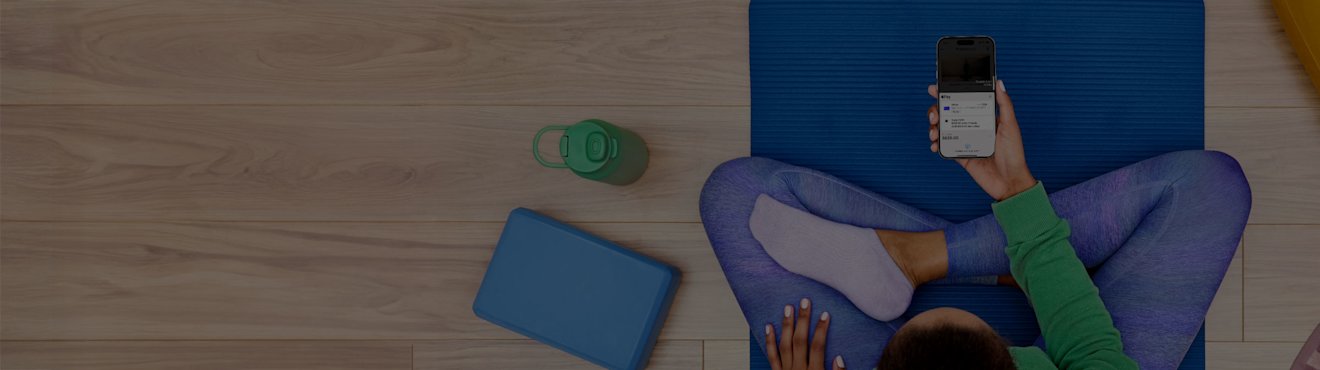 A person sitting cross-legged on a blue yoga mat holding a smartphone with a payment plan interface displayed, surrounded by a yoga bolster, water bottle, and other fitness accessories on a wooden floor.