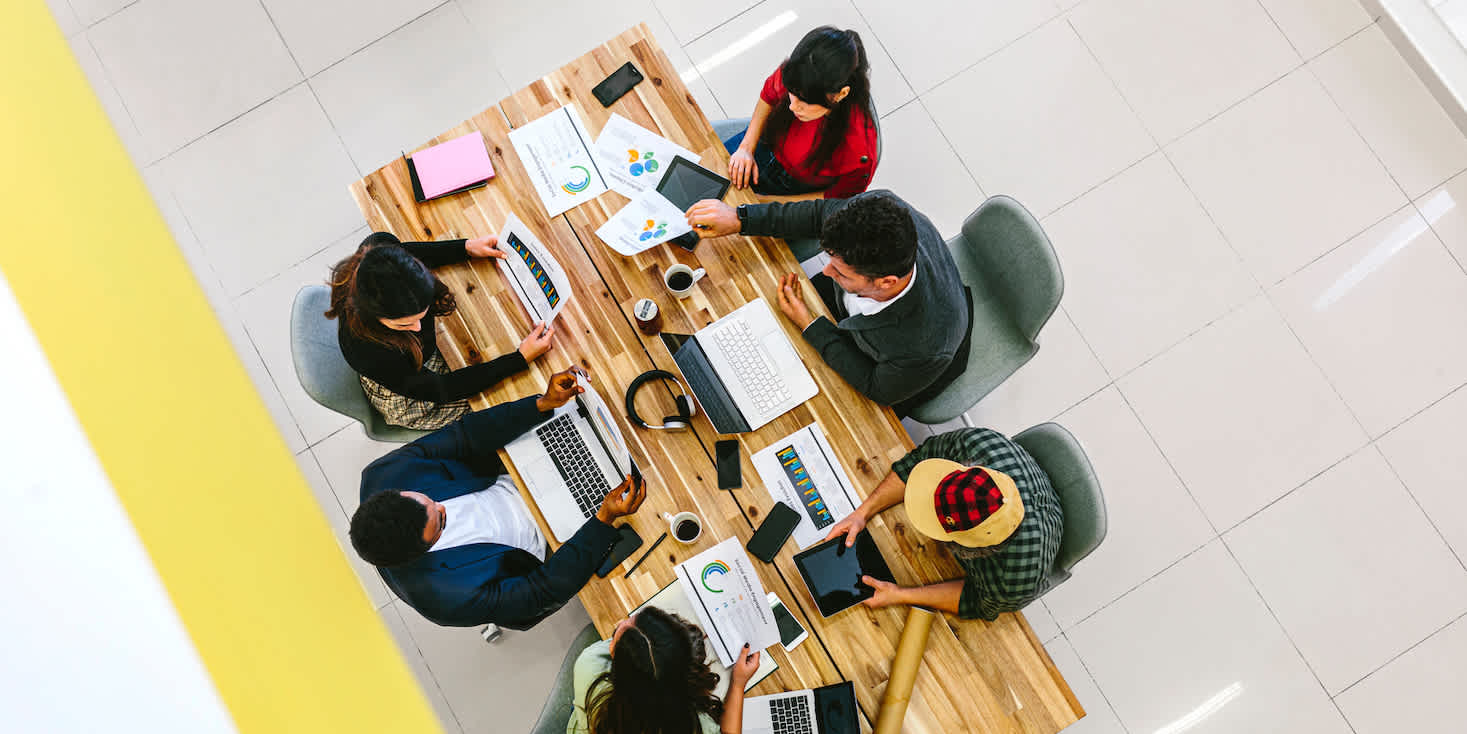 Overhead shot of young professionals around a wooden table having a meeting, with lots of papers and graphs and notepads that are helping with the plans.