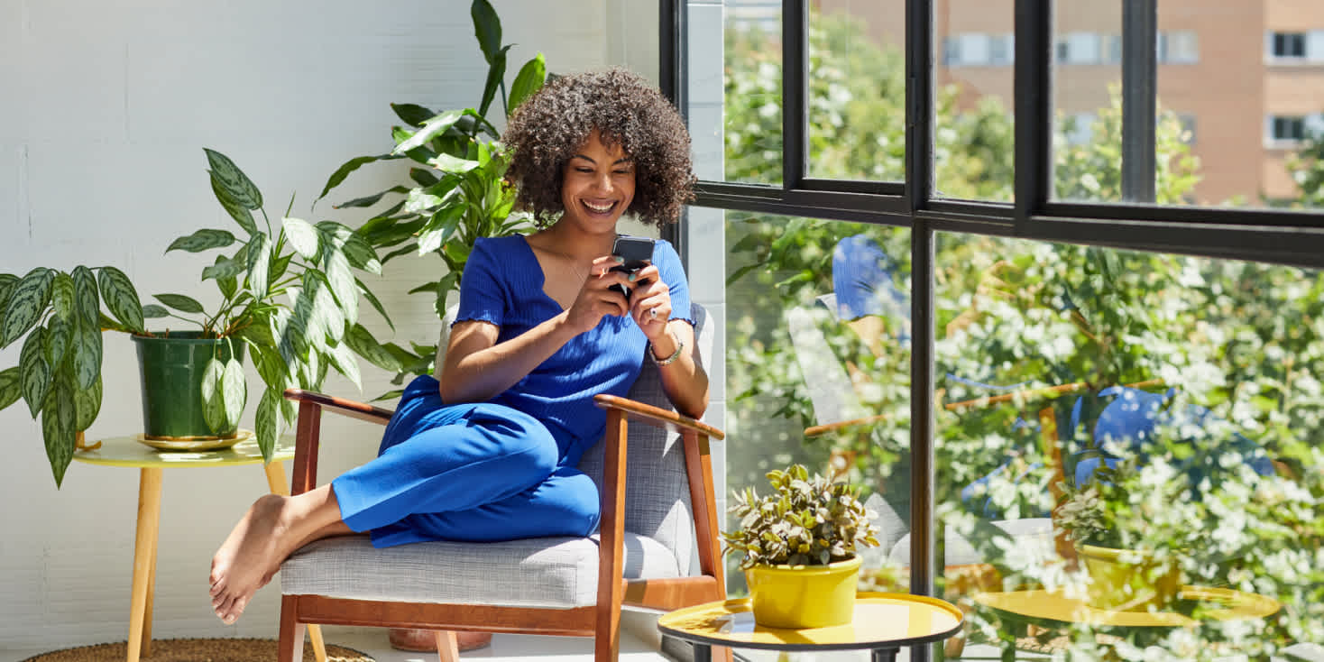 Woman sitting next to large window above trees, curled up on a chair and smiling as she makes a purchase on her phone